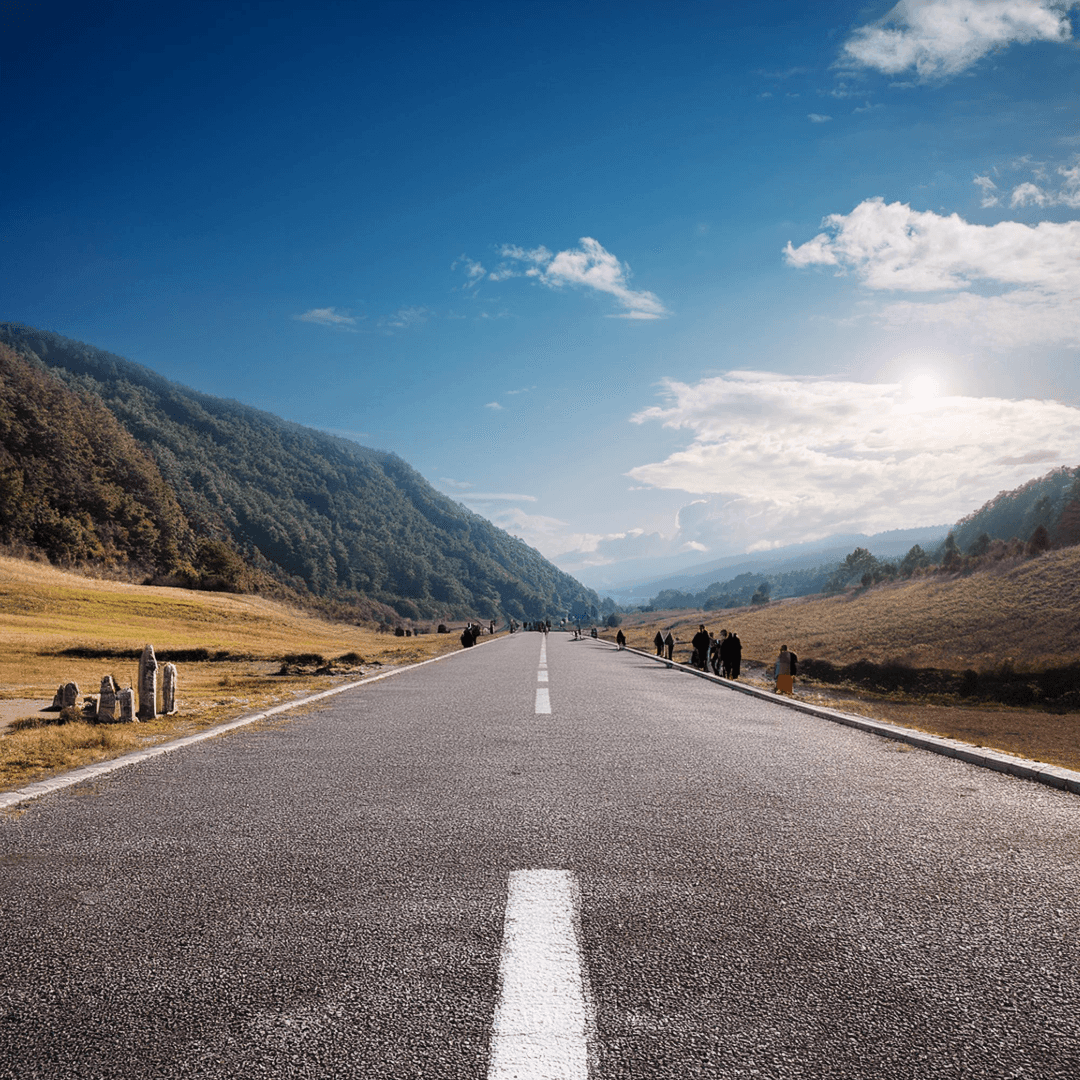 Straight road stretching towards distant hills under a blue sky, with people walking along the roadside.